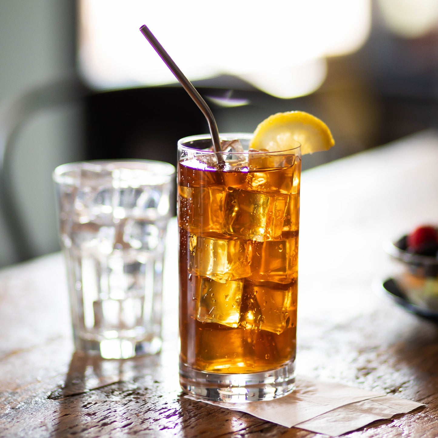A glass of Segafredo Zanetti Premium Iced Tea with a lemon wedge and a metal straw sits on a wooden table, next to a glass of water and a box of sachets.